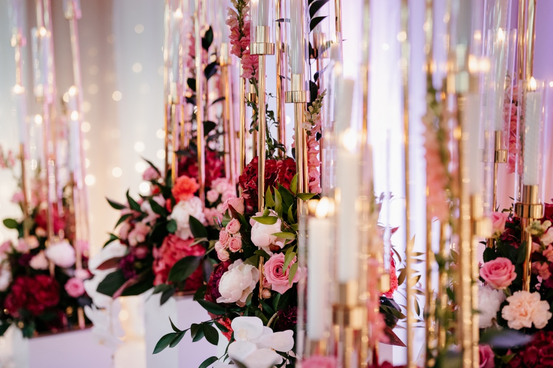 Piękne złote świeczniki z białymi świecami wśród różowo czerwonych żywych kwiatach.